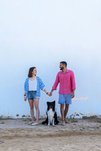 Happy couple standing with dog against wall while holding hands