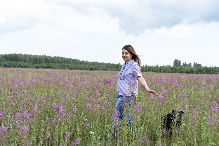 Happy young blond woman in  walking on field among flowers of fireweed with dog, travel with pets 