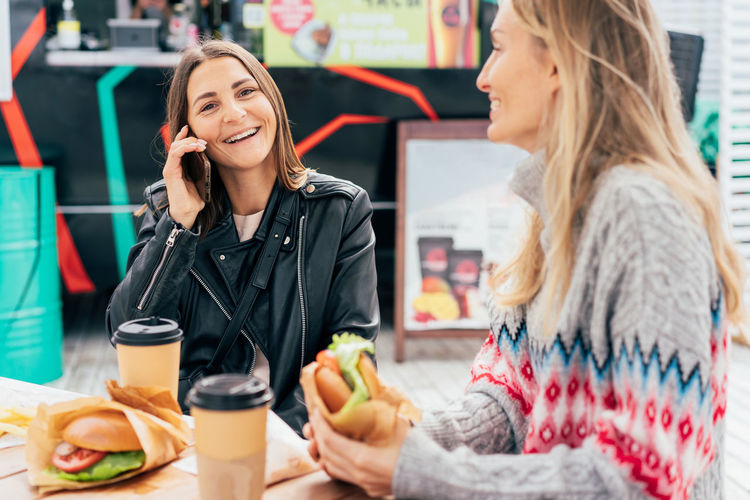 Two cheerful girlfriends are chatting and eating fast food at the street market.