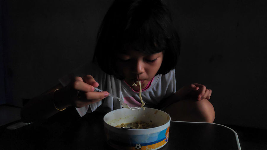Cute asian child girl eating delicious instant noodles with fork in the dark background