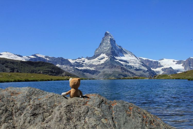 Stuffed teddy bear on rock formation by lake against clear blue sky
