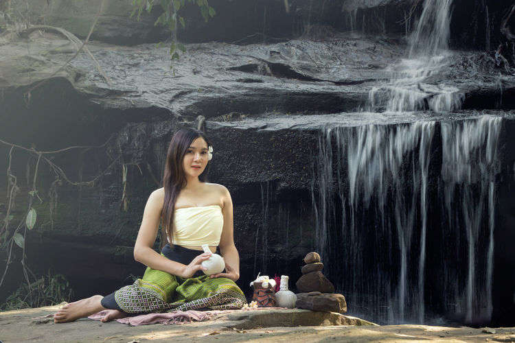 Portrait of smiling woman sitting on rock against waterfall