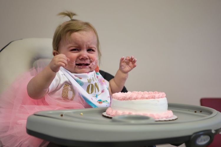 Cute baby girl crying while sitting with birthday cake on high chair at home