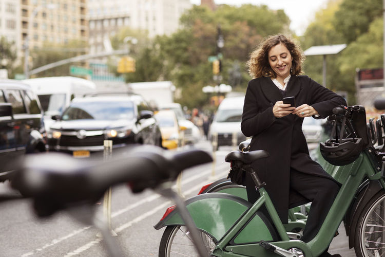 Smiling woman using mobile phone while standing by bicycle rack in city