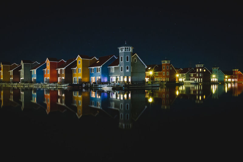Illuminated houses by river against sky at night