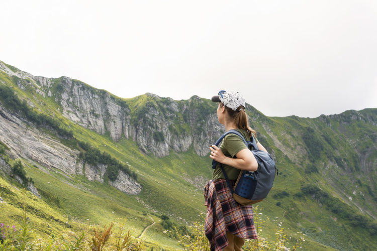 Young woman hiker in cap with backpack looking at mountain view in summer healthy active lifestyle