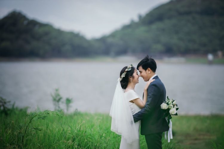Married couple embracing amidst grass against lake
