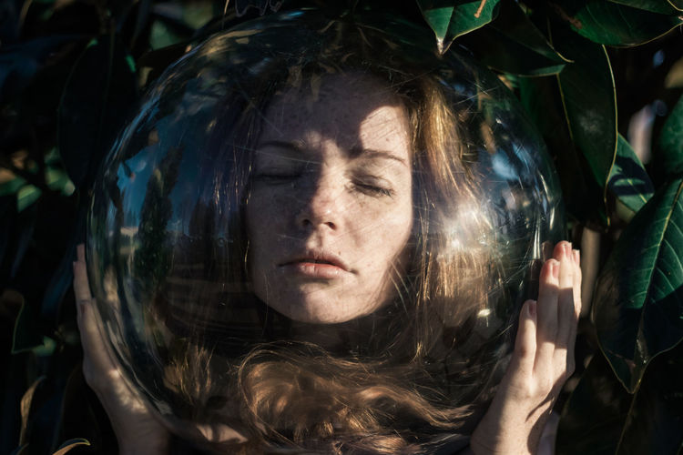 Close-up of young woman wearing glass ball on head against leaves