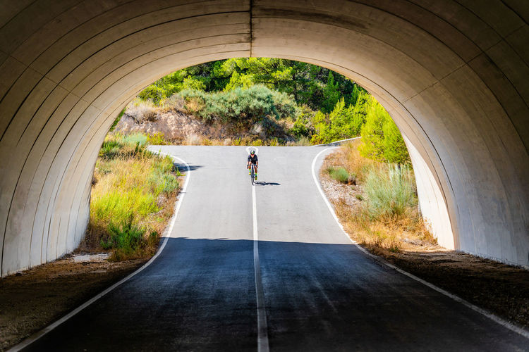 Full length of bicyclist in sportswear and helmet cycling on asphalt roadway under arched bridge in summer countryside
