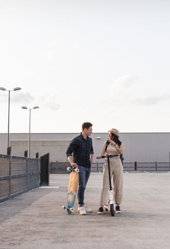 Young man and woman with longboard and electric scooter talking on parking deck
