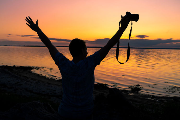 Silhouette woman photographing with arms raised while standing at beach during sunset