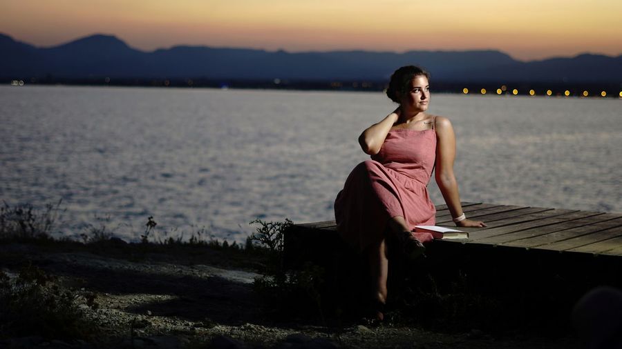 Portrait of woman sitting on shore against sky during sunset