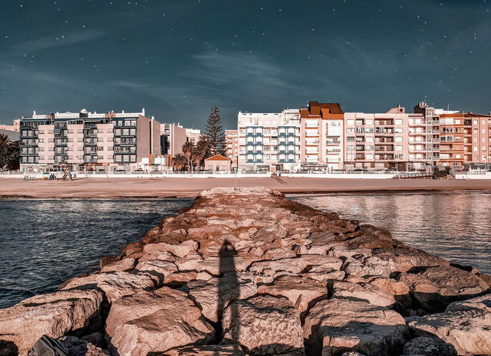 Buildings by sea against sky in city and a person's shadow on the rocks