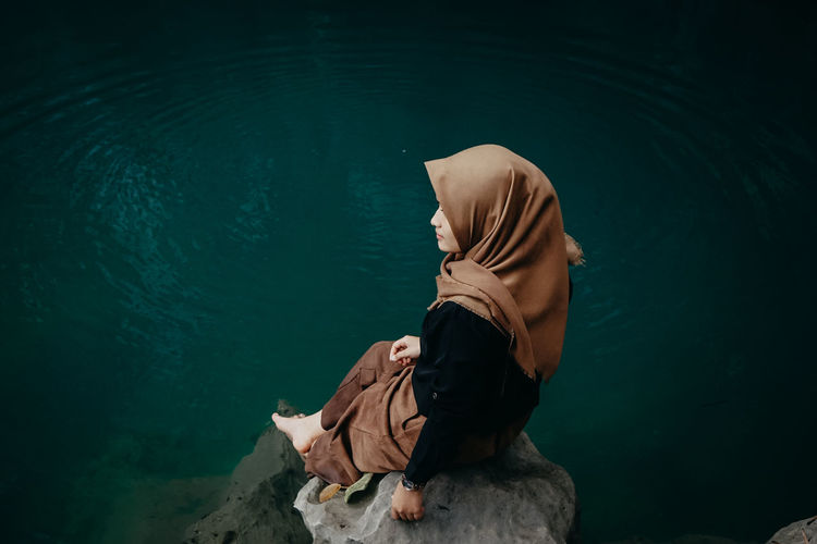 High angle view of woman wearing hijab sitting on rock by lake