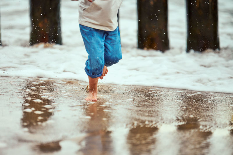 Little boy in blue pants running on sea coast and playing with sea waves, unfocused blurred legs