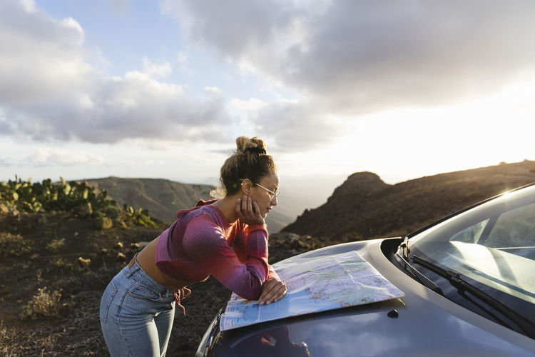 Young woman leaning on car hood checking map at sunset