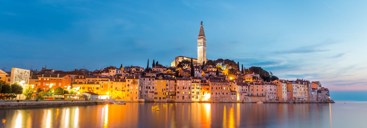 Illuminated buildings by sea against sky in city