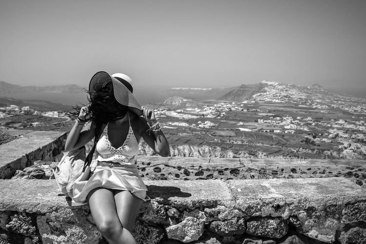 Woman with hat and tousled hair sitting on retaining wall against sky during sunny day