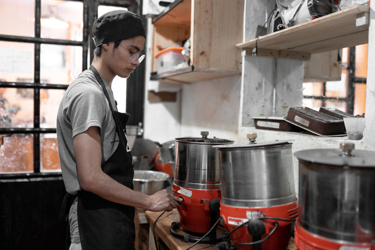 Side view of hispanic young man in apron and cap turning on chocolate tempering machine while working in kitchen of local production in chiapas, mexico