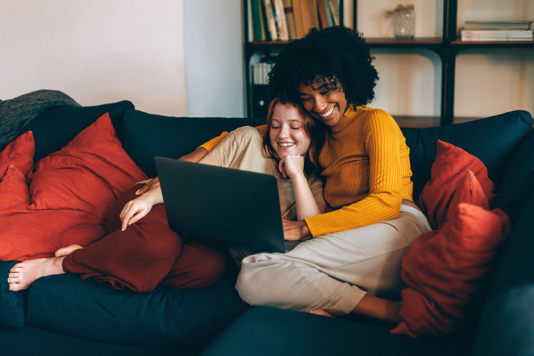 Multiethnic female relaxed lesbian couple on couch watching laptop and bonding