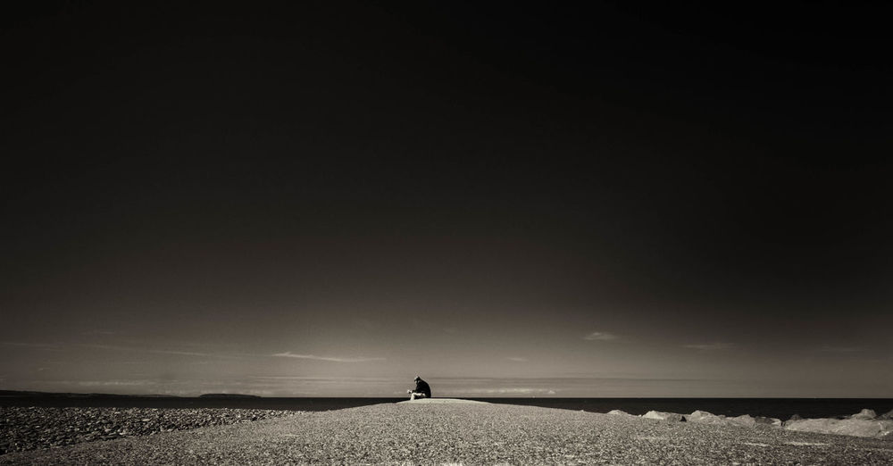 Distant view of man sitting by sea shore against sky
