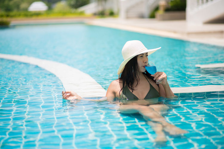 Young woman wearing hat drinking juice while relaxing in swimming pool 