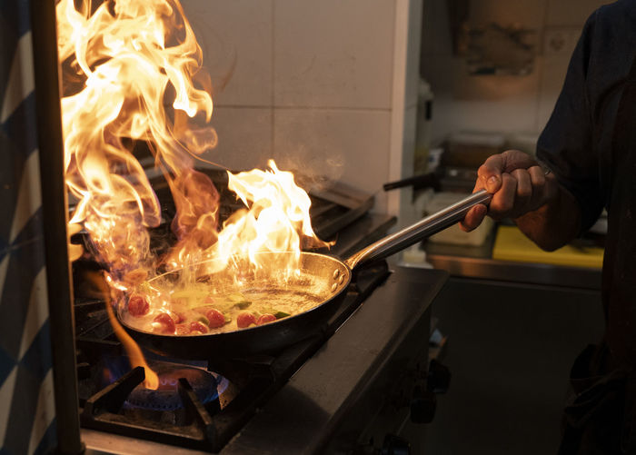 Crop anonymous cook holding frying pan with burning flame while preparing meal with flambe in restaurant kitchen