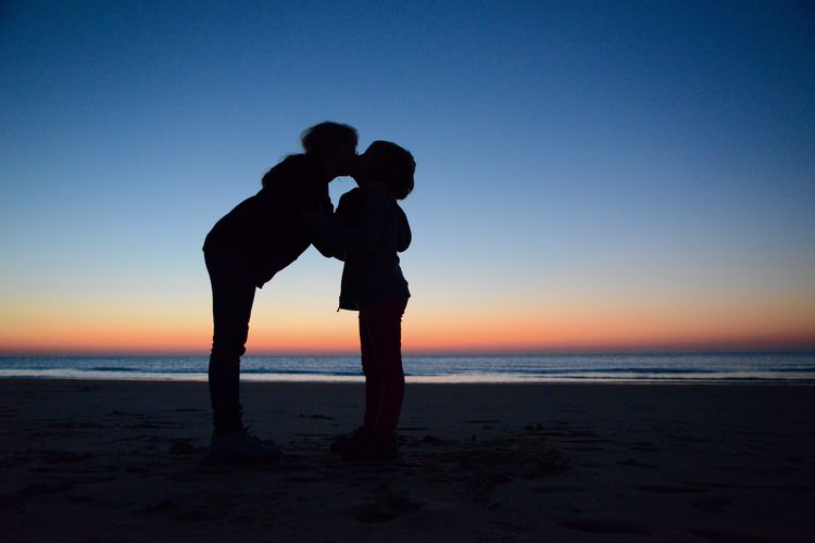 Silhouette siblings embracing while standing at beach against sky during sunset