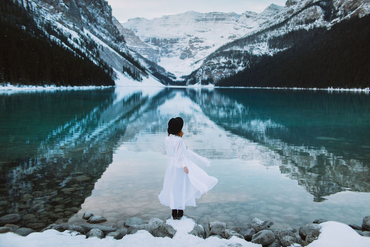 Back view of anonymous female in white dress standing towards clean water of lake louise against snowy mountain ridge on winter day in alberta, canada