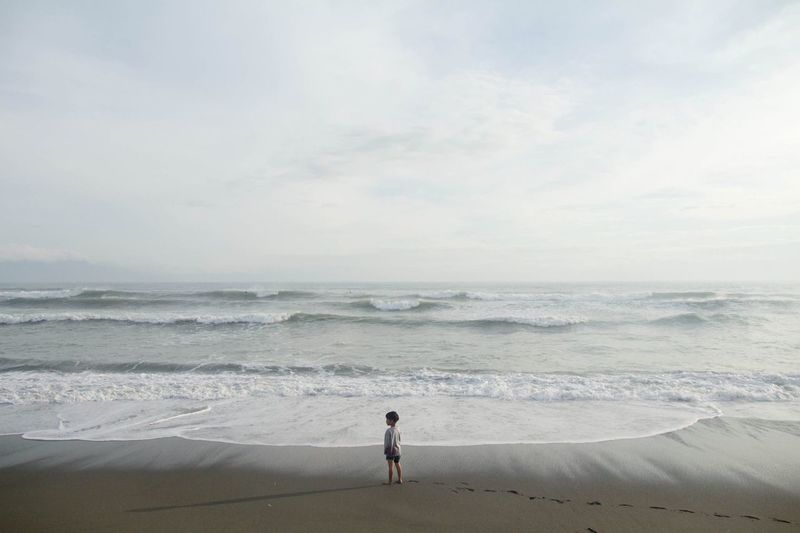 Rear view of boy standing at beach against sky