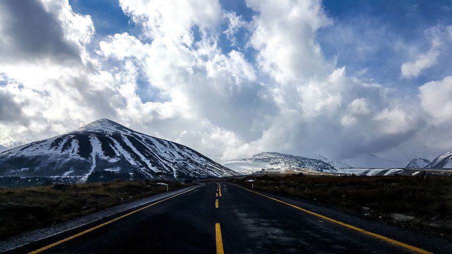 Empty road leading towards snow covered mountains against cloudy sky