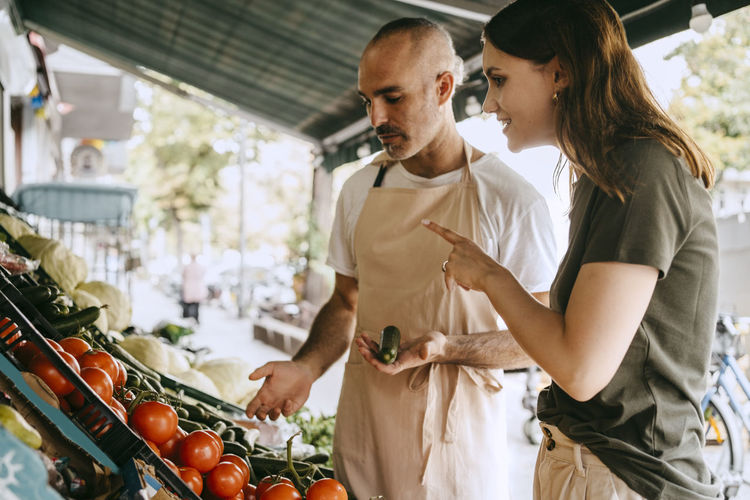 Side view of female customer gesturing while talking to male vendor holding vegetable at market