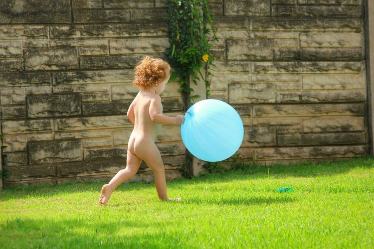 Full length of naked girl holding blue balloon while running on grass