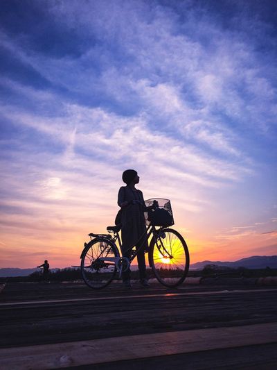 Silhouette of woman on bicycle against cloudy sky at sunset
