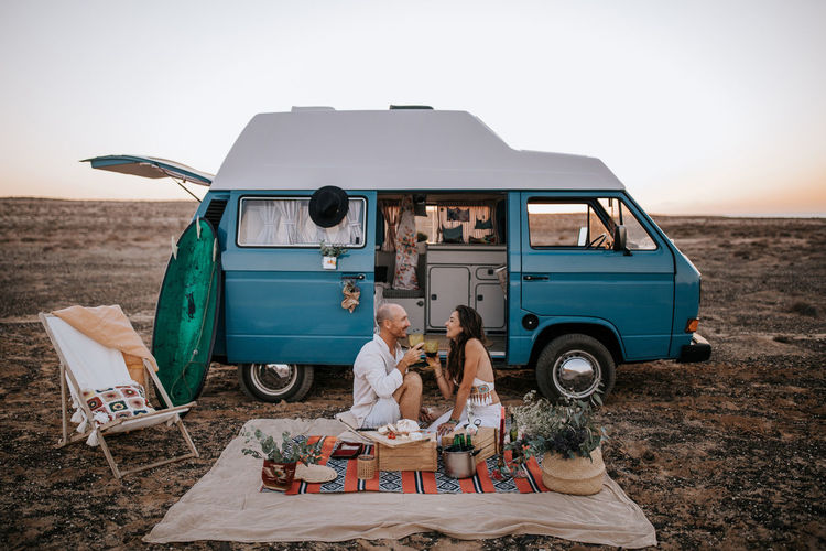 Side view of traveling couple sitting on blanket near van in savanna and having picnic while enjoying summer vacation and looking at each other
