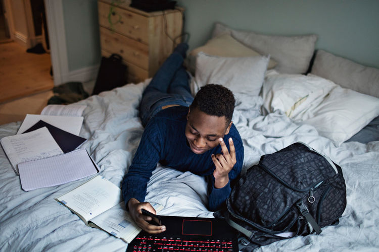 Smiling teenage boy using social media while doing homework on bed at home