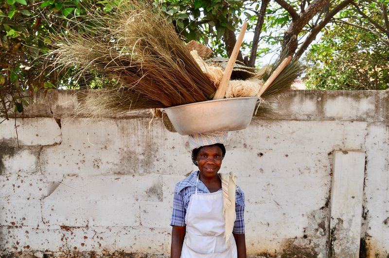 Portrait of smiling woman carrying basket on head while standing against wall