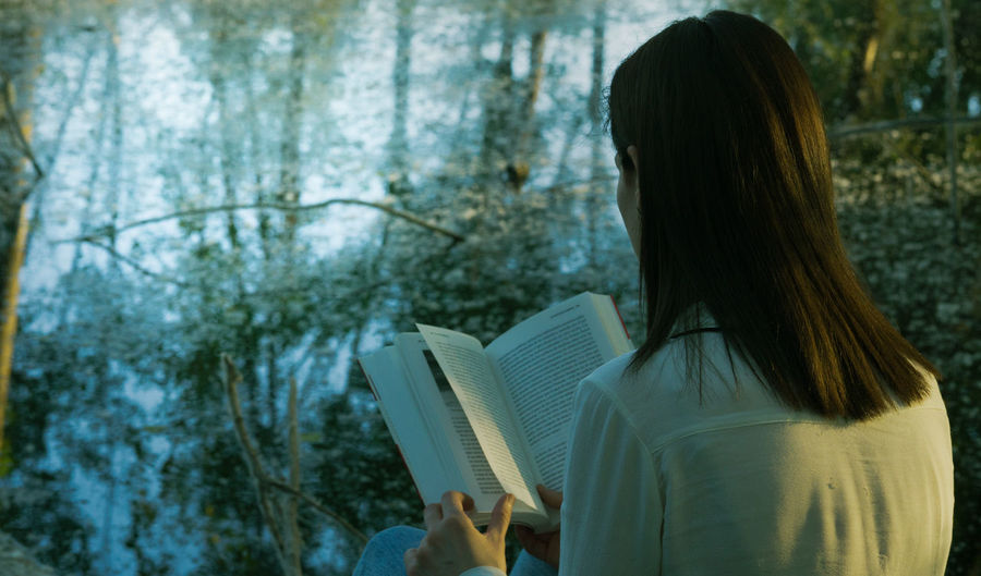Rear view of woman reading book while sitting at lakeshore