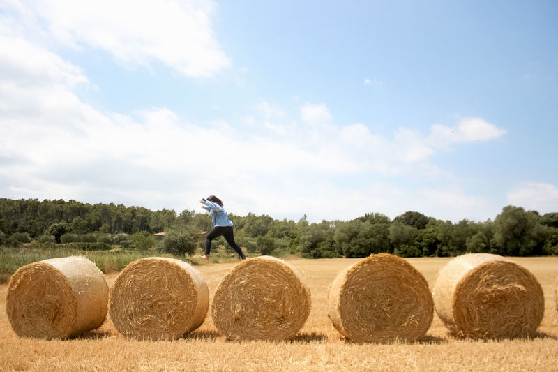 Side view of woman jumping on hay bales at field against sky