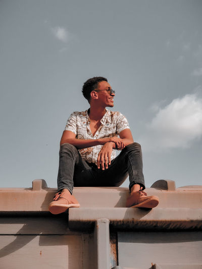 Young man looking away while sitting on wall against sky
