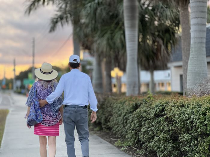 Senior couple walking slowly at sunrise on palm tree lined sidewalk against sky.  rearview portrait.