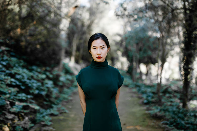 Young ethnic female in trendy dress on in green bushes and looking at camera in aged garden