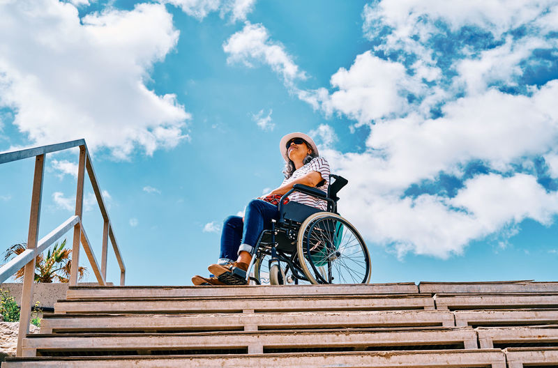 Low angle full body of positive handicapped female sitting in wheelchair near stairway looking away against blue sky in city