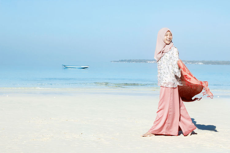 Side view of smiling young woman wearing hijab walking at beach against clear blue sky during sunny day