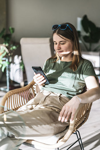 Young pretty woman in green beige clothes sitting in wicker chair in sunlight using smartphone