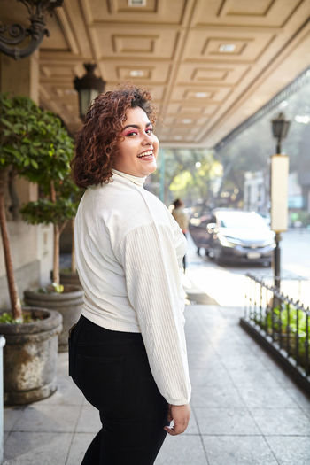 Side view of plump female wearing trendy clothes standing in city on sunny day and confidently looking at camera
