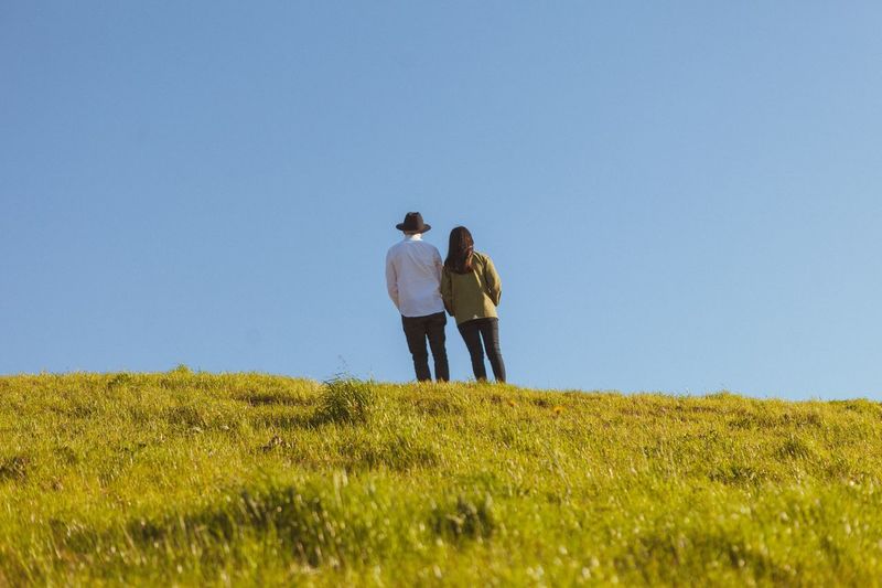 Rear view of man and woman walking on grassy field against clear sky