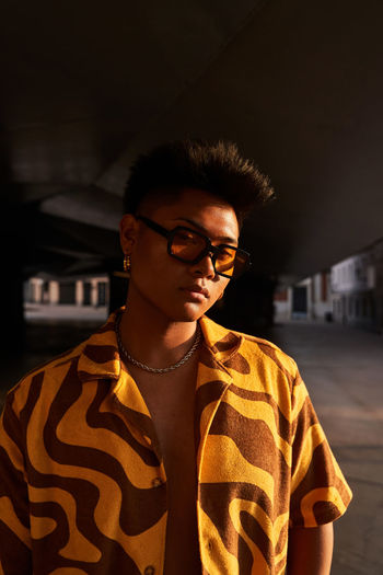 Confident asian male wearing cool shirt with sunglasses and accessories looking at camera while standing in sundown light on street