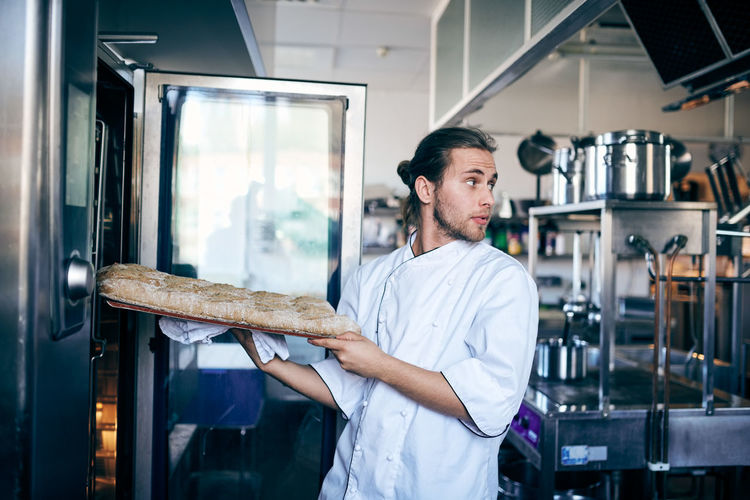 Male chef putting bread in oven while looking away at commercial kitchen