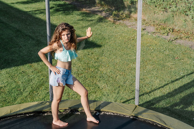 Horizontal photo of young brunette teenager on a trampoline leaning with her hand on the net on a park. the girl wears short jeans and a top and looks tired, passive and without energy.